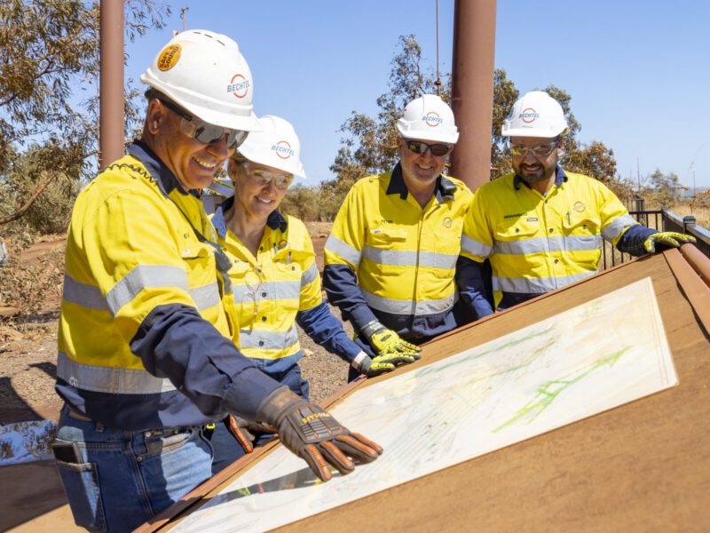 Four construction workers in yellow safety gear and helmets review plans on a large outdoor table at a work site where Bechtel was selected for preparatory engineering work at the Oklahoma smelter.