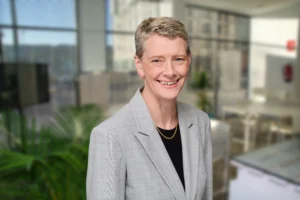 A person with short hair wearing a light gray suit jacket and black top stands in a modern, bright office setting, smiling at the camera.
