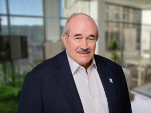 An older man with a mustache and receding hairline wearing a dark blazer and light shirt stands in a bright office setting.