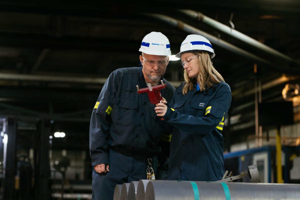 Two workers in protective gear use a measuring tool to inspect cylindrical metal rods in an industrial facility.