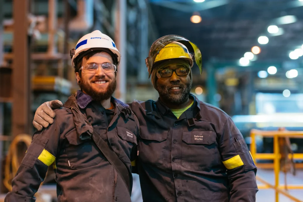 Two workers in PPE clasp shoulders and smile in front of several large machines in a warehouse.