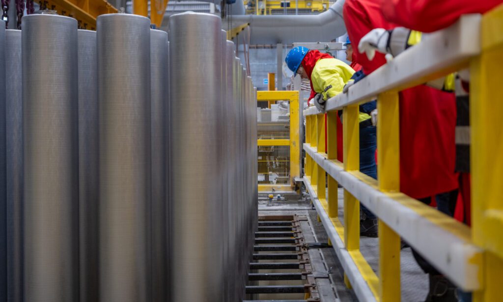 Workers in safety gear stand on a platform beside large metal cylinders in an industrial facility, with yellow railings and equipment visible.