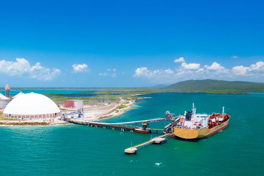 A large cargo ship is docked at an industrial port with a white dome storage facility by the coast, under a clear blue sky.