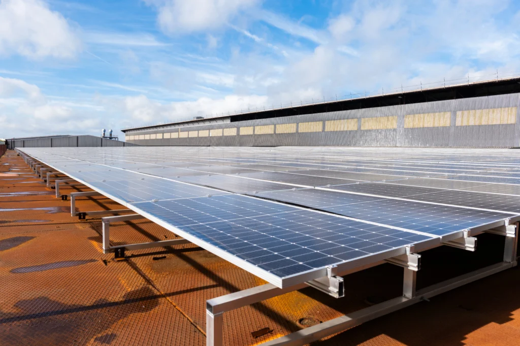 Rows of solar panels installed on a rust-colored metal rooftop under a partly cloudy sky.