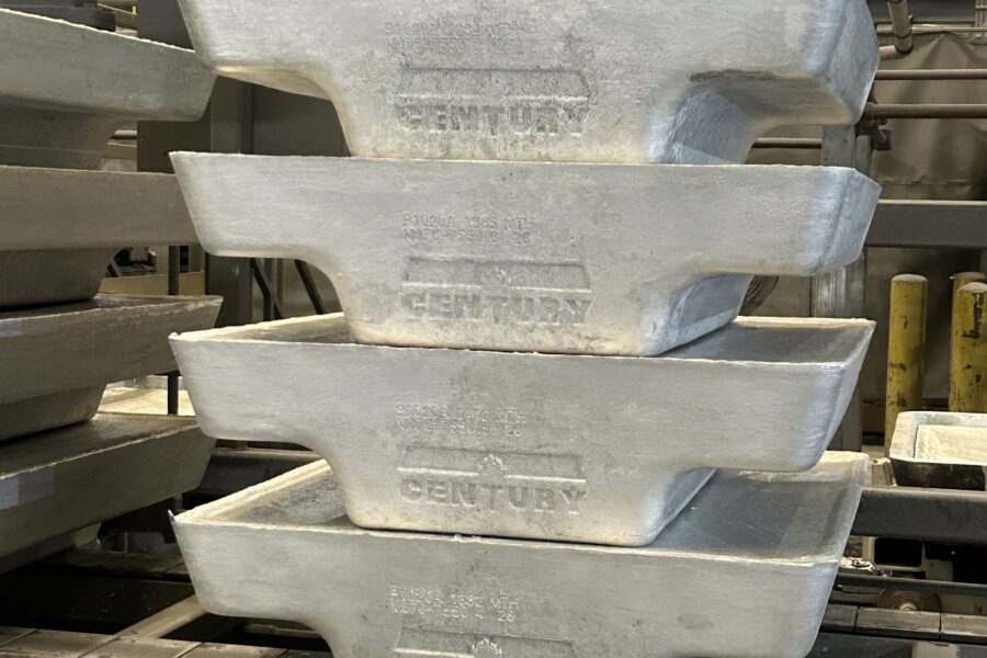 Four large metal ingots labeled "CENTURY" are stacked in an industrial warehouse setting with machinery and overhead lights visible in the background.