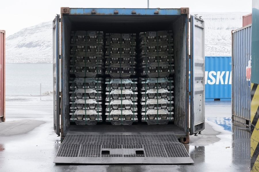 A shipping container with its doors open, filled with stacked metal ingots, is positioned on a wet outdoor surface near water and other containers.