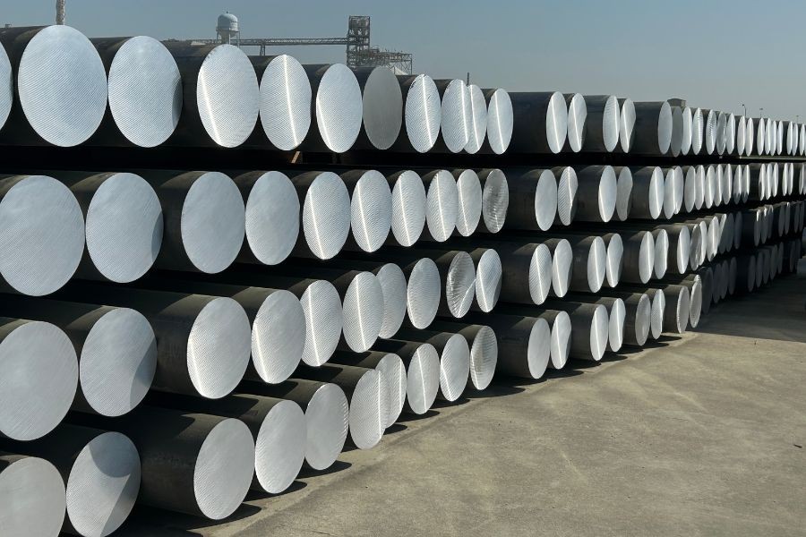 Stacks of large cylindrical metal billets are arranged in rows outdoors on a concrete surface under clear sky.