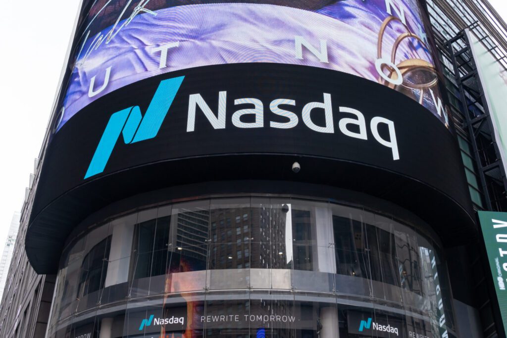 Large electronic Nasdaq sign displayed on the exterior of a building in Times Square, New York City, with partial reflections of nearby buildings visible in the glass.