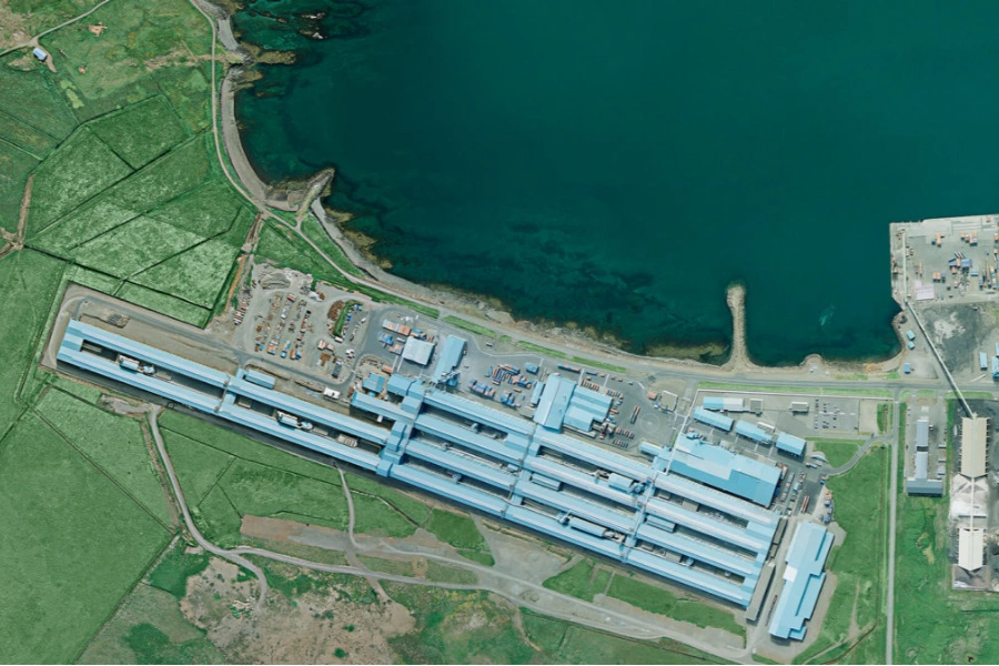 Aerial view of a large industrial facility with blue rooftops situated next to a body of water and surrounded by green fields.