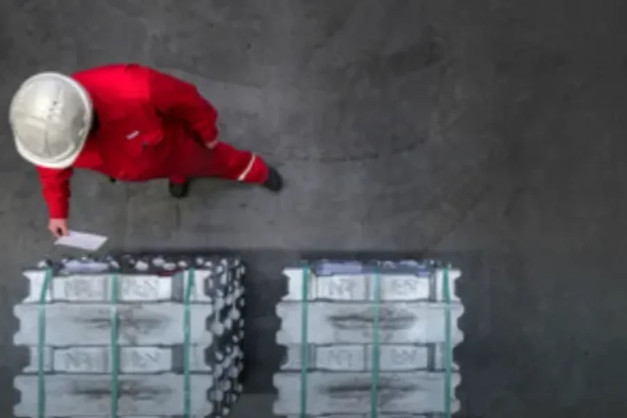 A person in a red uniform and helmet stands next to two stacks of metal ingots on a warehouse floor, holding a piece of paper.