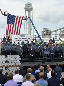 A group of workers and officials stand on a stage with an American flag and a "Make Aluminum Great Again" sign at an outdoor event; aluminum pipes are stacked in the foreground.