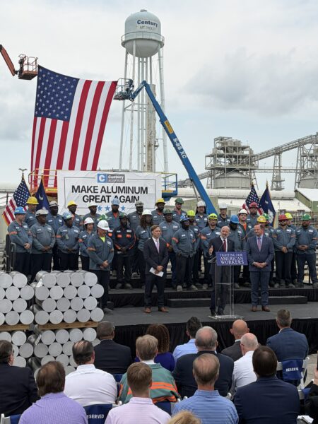 A group of workers and officials stand on a stage with an American flag and a "Make Aluminum Great Again" sign at an outdoor event; aluminum pipes are stacked in the foreground.