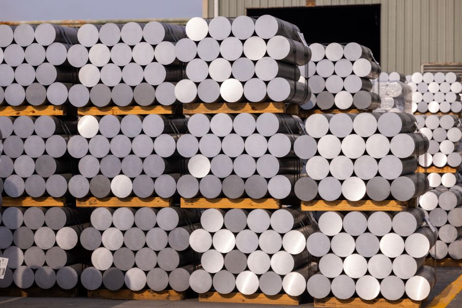 Stacks of cylindrical metal billets are arranged on wooden pallets outside a warehouse building.